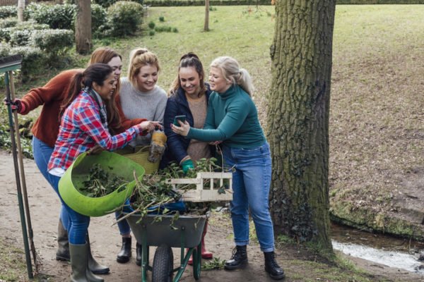 A diverse group of women holding gardening equipment filled with garden debris, looking at a mobile phone and laughing
