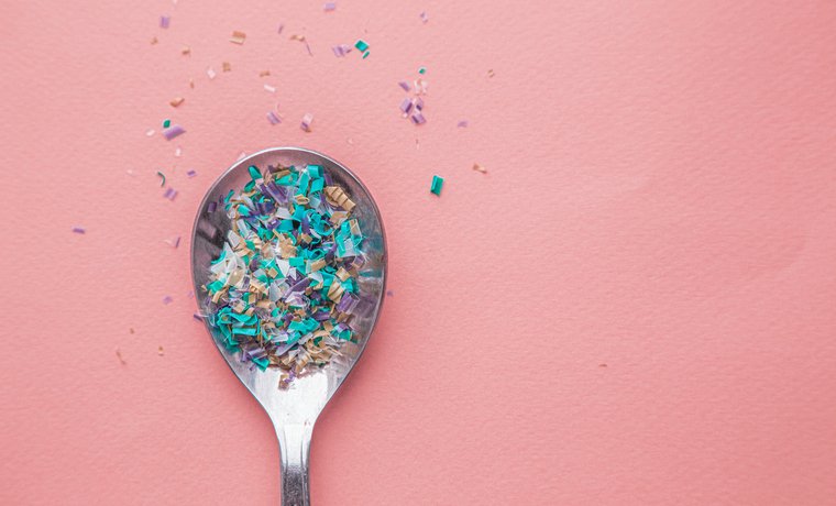 A spoon full of tiny Microplastics particles, viewed from above against a pink background