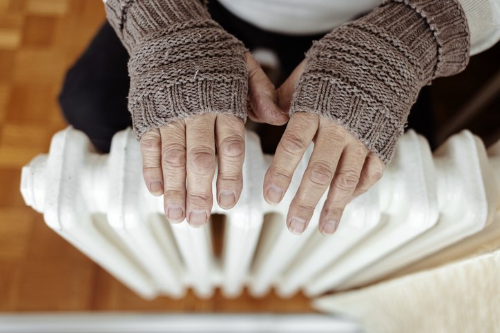 Old man's hands in knitted gloves on heating radiator at home during the day