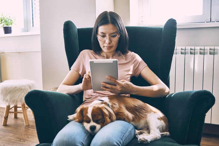 Young woman at home sitting in a chair using her tablet, with her puppy lying down in her lap