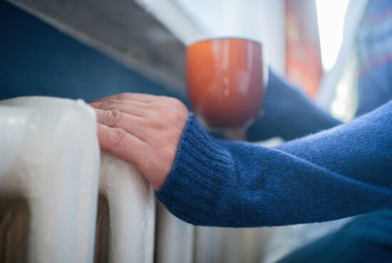 Man in warm sweater with hot tea near radiator in winter