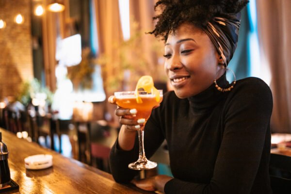 Side view portrait of an African woman drinking cocktails, sitting by bar in a modern and luxurious restaurant at night