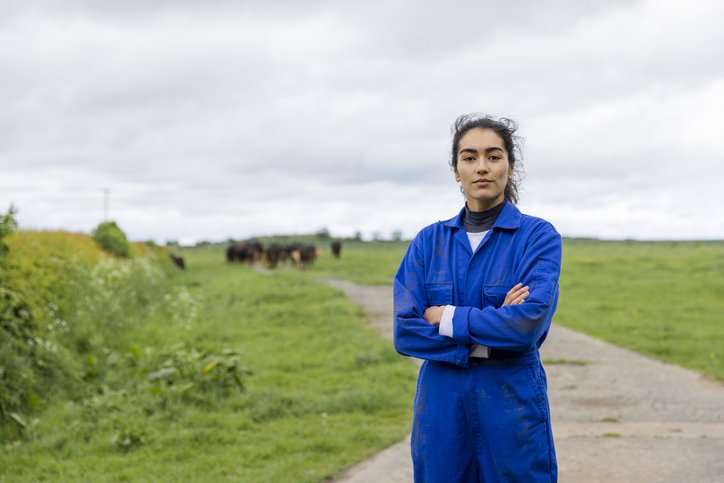 A portrait of a young female farmer wearing overalls, standing in an agricultural field at the farm she works at in Embleton, North East England while looking at the camera with a serious look on her face and her arms crossed