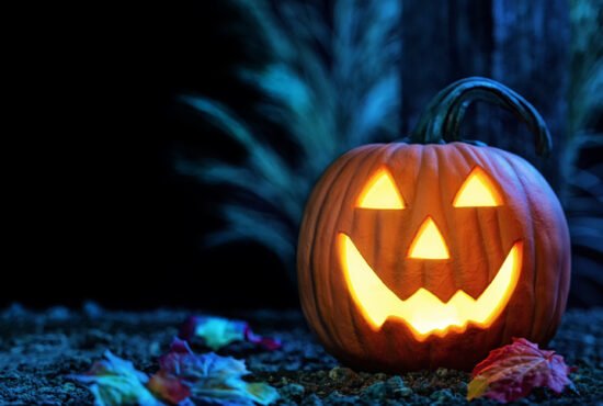 A smiling Jack O’ Lantern carved from a pumpkin looking at the camera, glowing from within, sitting on the dirt with autumn leaves in front of a wooden post at light lit from the moonlight