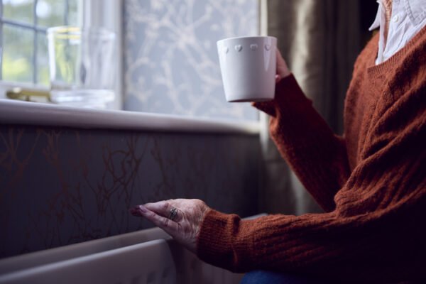 Senior woman with hot drink trying to keep warm by radiator at home in cost of living energy crisis
