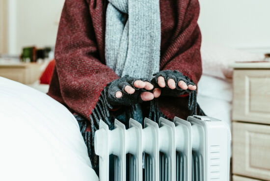 A retired senior man in his 70s sits at home inside his cold house in winter. It is so cold that he is wrapped up in warm winter clothing, including gloves and blanket, and is holding his hands over an electric heater for some extra warmth and comfort.