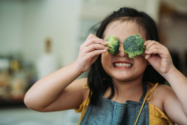Young girl smiling in the kitchen, holding broccoli florets over her eyes