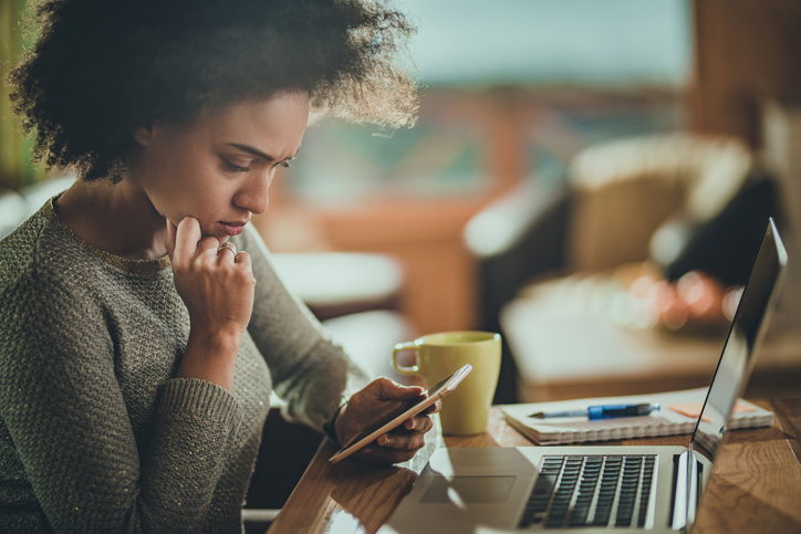 Young black woman reading a problematic text message on her mobile phone while working at home